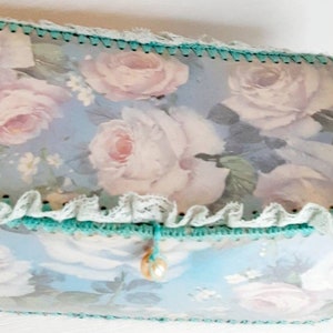 May include: A light blue box with a floral design featuring white roses and green trim. The box has a white lace trim and a pearl-like button.