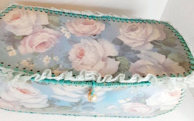 May include: A light blue box with a floral design featuring white roses and green trim. The box has a white lace trim and a pearl-like button.