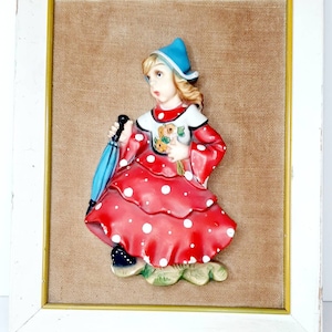 May include: A framed 3D wall art piece featuring a girl in a red and white polka dot dress, blue hat, and holding a blue umbrella. The background is a brown burlap fabric.