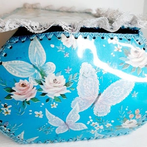 Pu&ograve; includere: Una scatola ovale blu con un bordo di pizzo bianco. La scatola &egrave; decorata con un motivo floreale con rose bianche e farfalle.