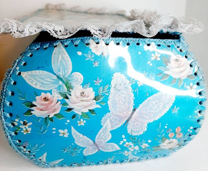 Pu&ograve; includere: Una scatola ovale blu con un bordo di pizzo bianco. La scatola &egrave; decorata con un motivo floreale con rose bianche e farfalle.