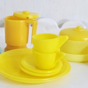 Op de afbeelding: Een set gele plastic speelgoedkeukengerei, inclusief een theepot, een pan, een koekenpan, een bord, een kom en twee kopjes. De set is perfect voor rollenspel.