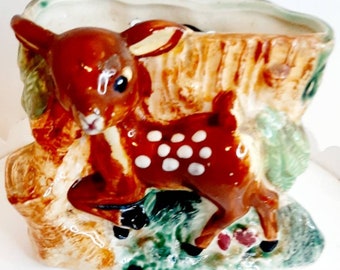 May include: A ceramic planter shaped like a fawn with brown and white spots, standing on a tree stump with green leaves. The planter is a light brown color with a white glaze.