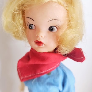 K&ouml;nnte beinhalten: Eine Vintage-Puppe mit blondem Haar, das zu einem Pferdeschwanz und Locken gestylt ist. Die Puppe hat einen roten Schal, ein blaues Kleid und einen wei&szlig;en G&uuml;rtel. Das Gesicht der Puppe hat bemalte Merkmale, einschlie&szlig;lich roter Lippen und gro&szlig;er Augen.