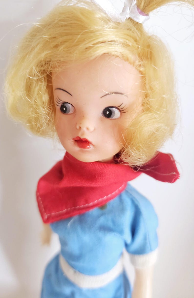 K&ouml;nnte beinhalten: Eine Vintage-Puppe mit blondem Haar, das zu einem Pferdeschwanz und Locken gestylt ist. Die Puppe hat einen roten Schal, ein blaues Kleid und einen wei&szlig;en G&uuml;rtel. Das Gesicht der Puppe hat bemalte Merkmale, einschlie&szlig;lich roter Lippen und gro&szlig;er Augen.