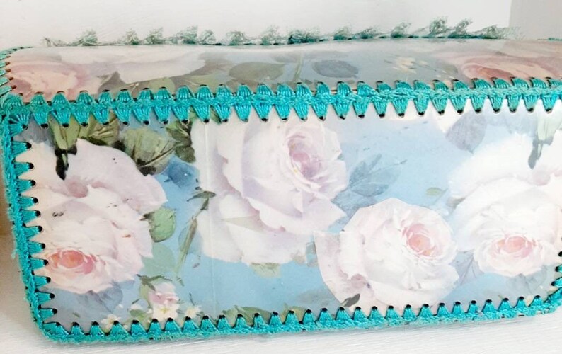 May include: A turquoise blue crocheted trim surrounds a light blue background with a floral pattern of white roses.