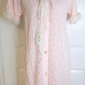 以下が含まれることがあります： 白いレースのトリムと白いリボンタイが付いたピンクのレースのナイトガウン。ナイトガウンはボタン留めになっており、サイズタグには「サイズ12 バスト34にフィット」と記載されています。