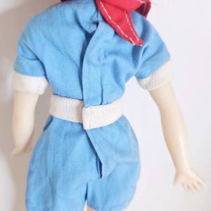 K&ouml;nnte beinhalten: Vintage-Puppe in einem hellblauen Spielanzug mit wei&szlig;en Verzierungen und passendem G&uuml;rtel. Die Puppe hat blondes Haar und einen roten Schal um den Hals gebunden. Die Arme der Puppe sind ausgestreckt.
