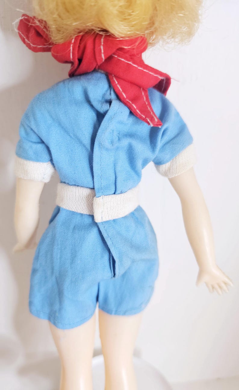 K&ouml;nnte beinhalten: Vintage-Puppe in einem hellblauen Spielanzug mit wei&szlig;en Verzierungen und passendem G&uuml;rtel. Die Puppe hat blondes Haar und einen roten Schal um den Hals gebunden. Die Arme der Puppe sind ausgestreckt.