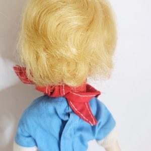 K&ouml;nnte beinhalten: Eine Vintage-Puppe mit blondem Haar, das mit einem wei&szlig;en Band zur&uuml;ckgebunden ist. Die Puppe tr&auml;gt ein blaues Outfit mit wei&szlig;en Verzierungen und einem roten Schal. Die Arme der Puppe sind sichtbar, vor einem wei&szlig;en Hintergrund.