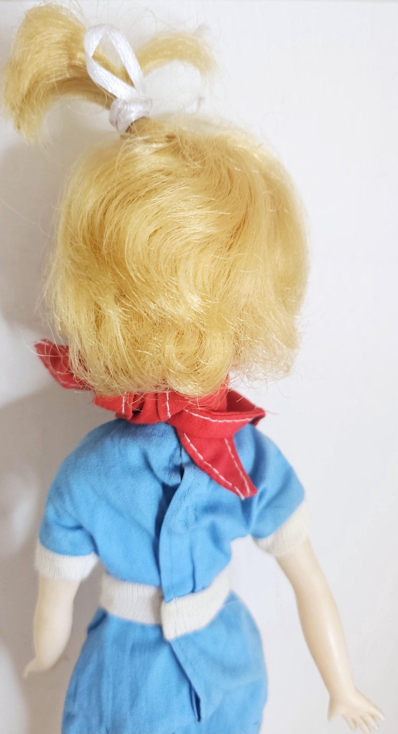 K&ouml;nnte beinhalten: Eine Vintage-Puppe mit blondem Haar, das mit einem wei&szlig;en Band zur&uuml;ckgebunden ist. Die Puppe tr&auml;gt ein blaues Outfit mit wei&szlig;en Verzierungen und einem roten Schal. Die Arme der Puppe sind sichtbar, vor einem wei&szlig;en Hintergrund.