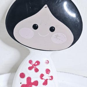 May include: A vintage-style doll-shaped clip. The doll has a black bob hairstyle, a light skin tone, and rosy cheeks. The white dress is adorned with pink flower designs. The clip is likely made of plastic or a similar material.