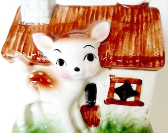 May include: A ceramic figurine of a white deer standing in front of a brown and white house with a white bird on the roof. The house has a brown roof, a chimney, and a window with a black star.