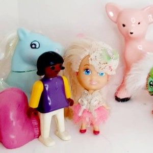 May include: A collection of vintage plastic toys including a pink deer, a blue horse, a green and pink rocking horse, a sleeping baby doll, a black doll wearing a purple shirt, and a blonde doll wearing a white dress and a pink tutu.