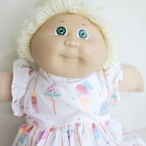 May include: A doll with a round vinyl face, green eyes, and light yellow yarn hair styled in a bun. The doll wears a white short-sleeved dress featuring a colourful pattern of ice cream cones, watermelon slices, and lollipops with small dots.