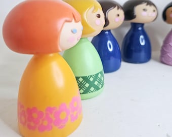 May include: Five colorful plastic dolls with painted faces and hair. The dolls are arranged in a row from left to right, with the doll on the left wearing a yellow dress with pink flowers, the next doll wearing a green dress with a plaid pattern, the next doll wearing a blue dress, the next doll wearing a blue dress, and the last doll wearing a pink dress.