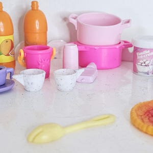 May include: A selection of miniature toy kitchen items in various colours. Includes orange juice bottles, a purple goblet, a teacup and saucer, a pink pot, a vanilla ice cream container, and a pizza.