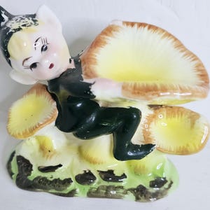 May include: A vintage ceramic figurine of an elf perched on a mushroom. The elf is wearing a dark green outfit and has a whimsical expression. The mushroom has a yellow and brown cap, and the base is green with dark spots.