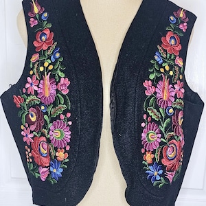 Könnte beinhalten: Schwarze Weste mit farbenfroher Blumenstickerei. Die Stickerei zeigt rote, rosa, blaue und gelbe Blumen mit grünen Blättern. Die Weste hat einen V-Ausschnitt und eine taillierte Passform, die ein traditionelles Volksdesign zeigt.