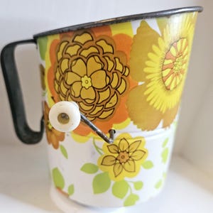 May include: Vintage metal kitchen utensil with a floral pattern in yellow, orange, and green. The utensil has a black handle and a white knob. The design features large flowers and leaves, giving it a retro aesthetic.