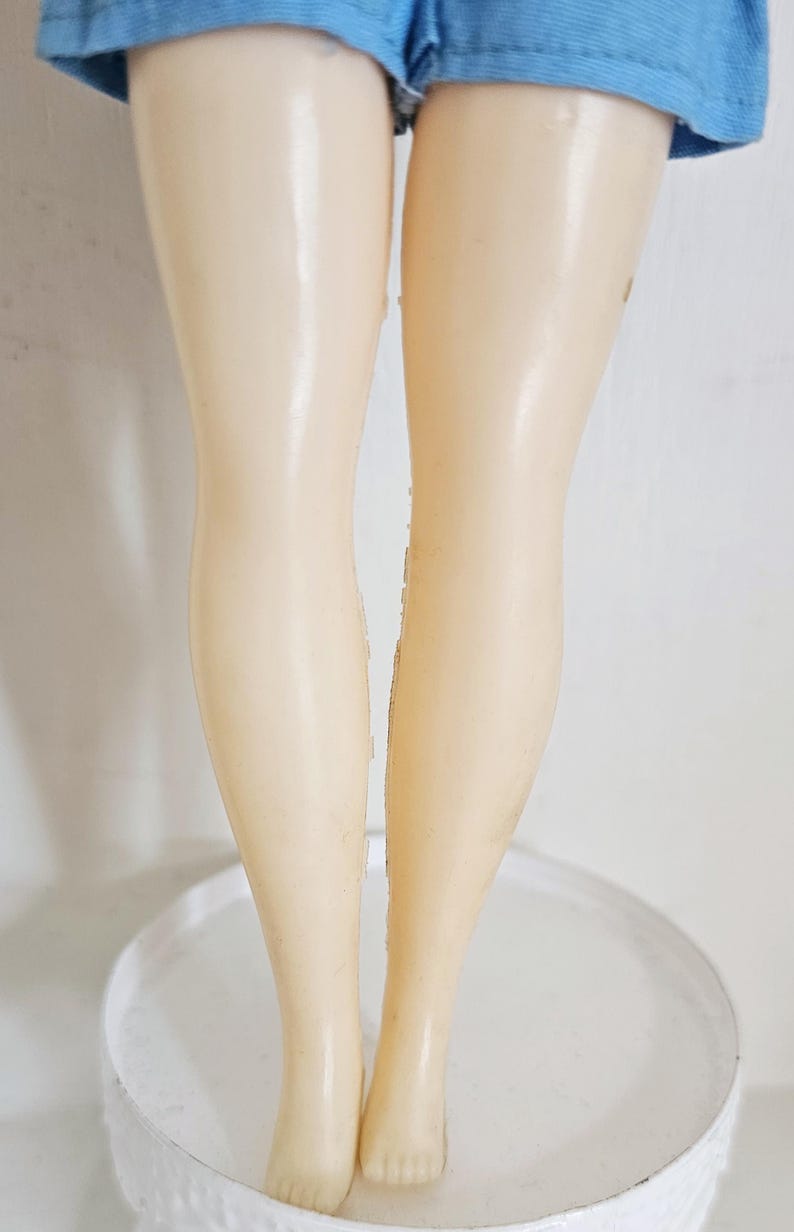 K&ouml;nnte beinhalten: Ein Paar Puppenbeine aus hellbeige Kunststoff, mit blauen Shorts oben sichtbar. Die Beine stehen auf einer wei&szlig;en, runden, strukturierten Oberfl&auml;che. Die Beine sind leicht an den Knien angewinkelt.