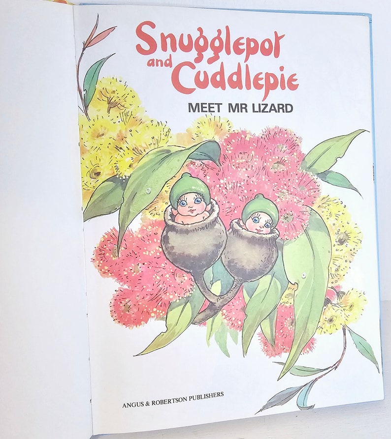 Puede incluir: Una portada de libro con dos peque&ntilde;as hadas con gorros verdes anidadas en nueces de goma. El t&iacute;tulo del libro es "Snugglepot and Cuddlepie Meet Mr Lizard" con un fondo de flores rosas y amarillas.