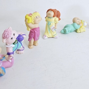 Puede incluir: Una colección de siete muñecas Cabbage Patch Kids, cada una con atuendos y accesorios únicos. Las muñecas están hechas de plástico y tienen rasgos pintados. Las muñecas están todas de pie o sentadas sobre una superficie blanca.