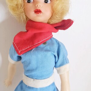 K&ouml;nnte beinhalten: Vintage Puppe mit blondem Haar, das mit einem wei&szlig;en Band zur&uuml;ckgebunden ist. Die Puppe tr&auml;gt ein blaues Outfit mit wei&szlig;en Verzierungen und einem roten Schal. Die Puppe hat bemalte Gesichtsz&uuml;ge, darunter rote Lippen und gro&szlig;e Augen.
