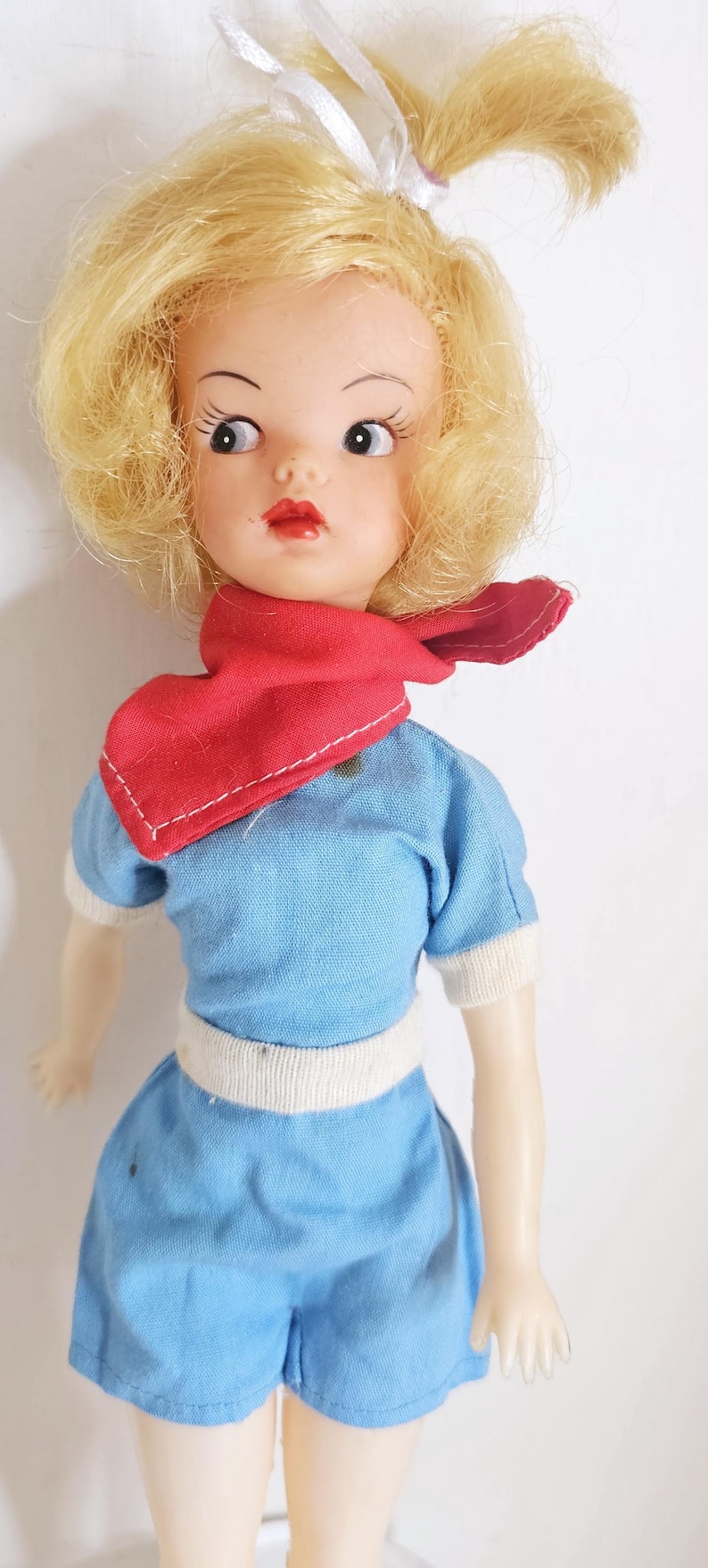 K&ouml;nnte beinhalten: Vintage Puppe mit blondem Haar, das mit einem wei&szlig;en Band zur&uuml;ckgebunden ist. Die Puppe tr&auml;gt ein blaues Outfit mit wei&szlig;en Verzierungen und einem roten Schal. Die Puppe hat bemalte Gesichtsz&uuml;ge, darunter rote Lippen und gro&szlig;e Augen.