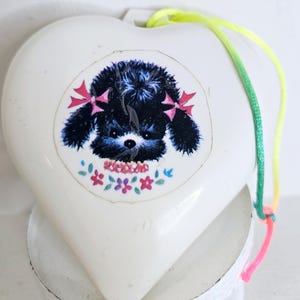 May include: A white heart-shaped ornament featuring a central graphic of a fluffy black dog with pink bows on its ears, surrounded by small colorful flowers. A bright neon yellow and pink cord is attached to the top for hanging purposes.