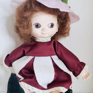 May include: A vintage doll with a flower-shaped hat and dress. The doll has brown curly hair, a burgundy dress with white petal-like accents, and green velvet shoes. The hat is white with a pink brim and green leaves.