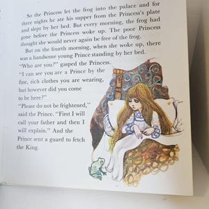 Puede incluir: Ilustraci&oacute;n de un libro infantil que representa a una princesa con un vestido blanco y pelo largo casta&ntilde;o. La princesa est&aacute; sentada en una cama con detalles dorados y azules ornamentados. Se ve una rana cerca de la cama. Tambi&eacute;n se ve texto del cuento.
