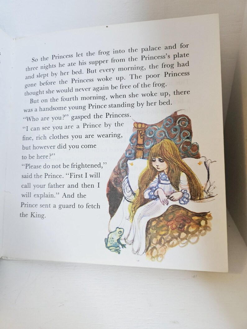 Puede incluir: Ilustraci&oacute;n de un libro infantil que representa a una princesa con un vestido blanco y pelo largo casta&ntilde;o. La princesa est&aacute; sentada en una cama con detalles dorados y azules ornamentados. Se ve una rana cerca de la cama. Tambi&eacute;n se ve texto del cuento.