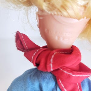 K&ouml;nnte beinhalten: Eine Vintage-Puppe mit blonden Haaren und einem roten Schal um den Hals. Die Puppe tr&auml;gt ein blaues Hemd. Das Gesicht der Puppe ist von der Kamera abgewandt.