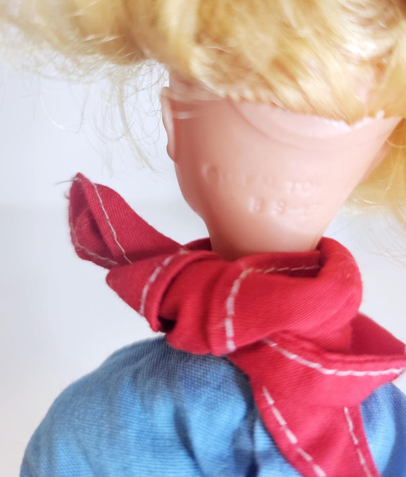 K&ouml;nnte beinhalten: Eine Vintage-Puppe mit blonden Haaren und einem roten Schal um den Hals. Die Puppe tr&auml;gt ein blaues Hemd. Das Gesicht der Puppe ist von der Kamera abgewandt.