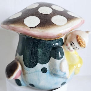 May include: A whimsical ceramic mushroom-shaped figurine with a brown cap dotted with white spots. The mushroom has a pink and blue base, and a small figure in yellow clothing is nestled against it. The figurine has a vintage aesthetic.