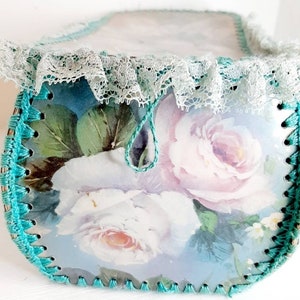 May include: A teal blue crocheted box with a white lace trim and a floral design featuring pink and white roses. The box has a teal blue drawstring.