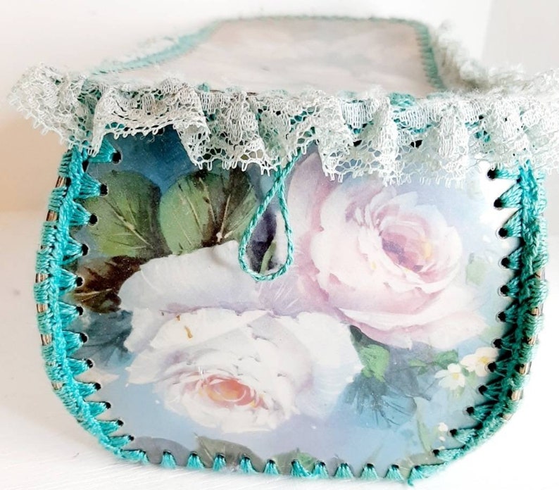 May include: A teal blue crocheted box with a white lace trim and a floral design featuring pink and white roses. The box has a teal blue drawstring.