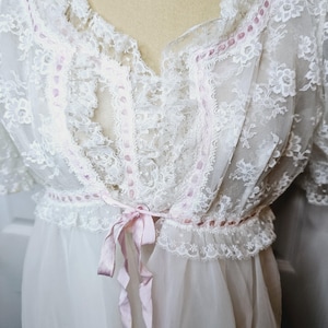 May include: A vintage white lace nightgown with short sleeves and a V-neckline. The gown features delicate lace detailing, a pink ribbon tie at the waist, and a flowing skirt. The intricate design and soft color palette evoke a romantic, classic aesthetic.