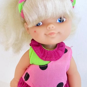 May include: A vintage doll with blonde hair and blue eyes. The doll is dressed in a pink and green outfit with black polka dots and a matching headband. A small red star is painted on the doll's cheek. The doll's outfit includes a ruffled skirt.