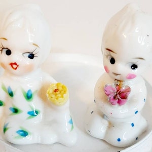 Può includere: Due figurine di porcellana di bambini, ciascuna con un fiore in mano. La bambina a sinistra strizza l'occhio e indossa un vestito floreale blu e verde. La bambina a destra tiene un fiore rosa e indossa un vestito bianco con pois blu.