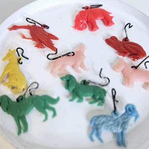 May include: A selection of plastic animal charms in various colours, such as red, yellow, and green. The charms, including an elephant and dogs, have metal loops for hanging. They are displayed on a white, textured surface.