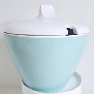 Può includere: Una zuccheriera azzurra con coperchio e manico bianchi. La ciotola ha una forma conica arrotondata e poggia su una base bianca. Il coperchio ha una piccola tacca per un cucchiaio.