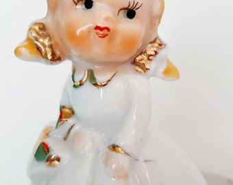 May include: A porcelain figurine of a girl with blonde hair, wearing a white dress with gold accents. She has a sweet expression on her face and is holding a small bouquet of flowers.