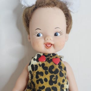 Puede incluir: Muñeca vintage con ojos azules, pecas y cabello castaño, vestida con un vestido con estampado de leopardo con dos acentos rojos en forma de corazón. Un lazo blanco adorna la cabeza de la muñeca. La muñeca tiene una expresión sonriente.
