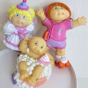 May include: Three Cabbage Patch Kids figurines. One is a girl wearing a white dress with pink trim and a pink hat. Another is a girl wearing a pink shirt and purple shorts. The third is a baby wearing a pink and white hat and sitting in a white tub.
