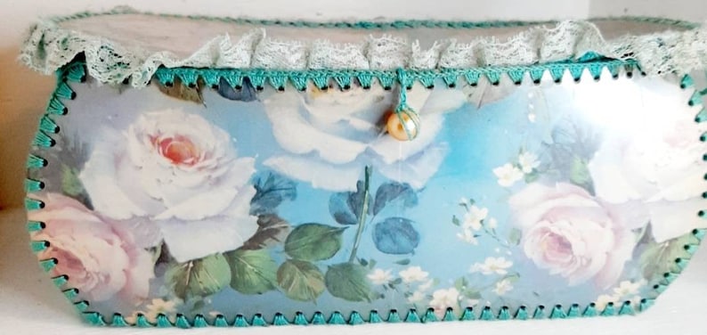 May include: A blue and white floral patterned box with a lace trim and a pearl button accent. The box is made of cardboard and has a scalloped edge.