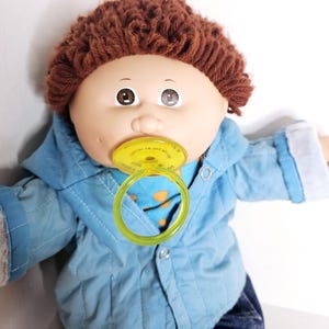 Op de afbeelding: Een pop met bruin garen haar en bruine ogen, gekleed in een lichtblauw doorgestikt jasje over een blauw shirt met oranje stippen. De pop heeft een gele plastic speen in de mond en draagt een donkerblauwe broek met zichtbare metalen drukknopen.