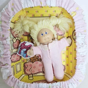 May include: A doll with blonde yarn hair in pigtails and blue eyes, wearing a light pink long-sleeved onesie with a lace collar. The doll is positioned inside a rectangular container lined with yellow wallpaper-patterned paper and a pink fabric border.