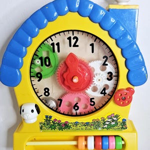 May include: A yellow and blue plastic toy clock with a white cat on top. The clock face has a clear cover with black numbers and colorful gears. A dog and a ladybug are on the clock. A colorful abacus is at the bottom.
